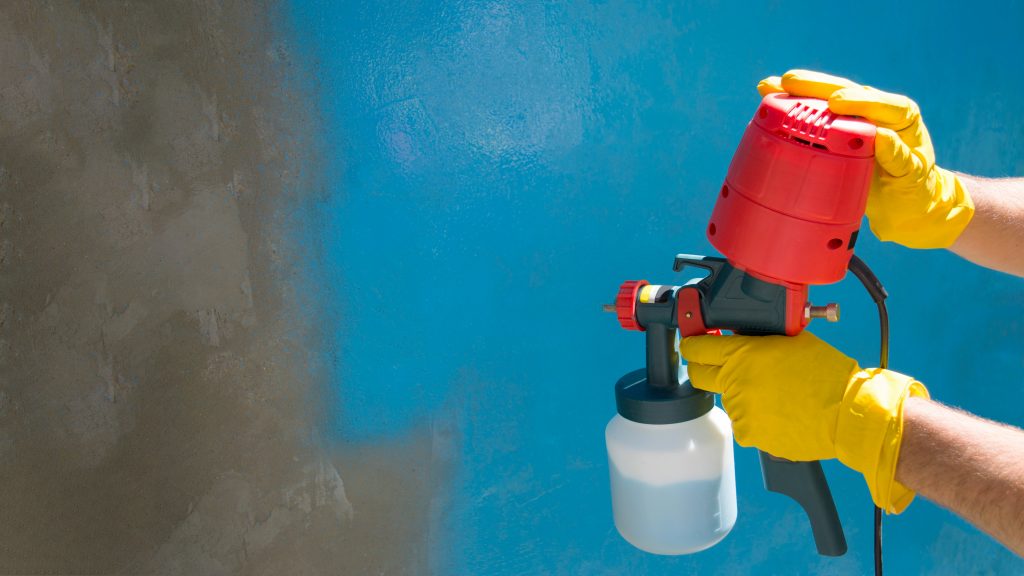 a workers hands in yellow gloves hold a hand held spray gun with an overhead compressor. Painting the wall of the house in blue