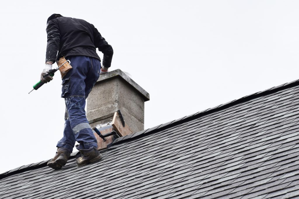 Roofer construction worker repairing chimney on grey slate shingles roof of domestic house, sky background with copy space.