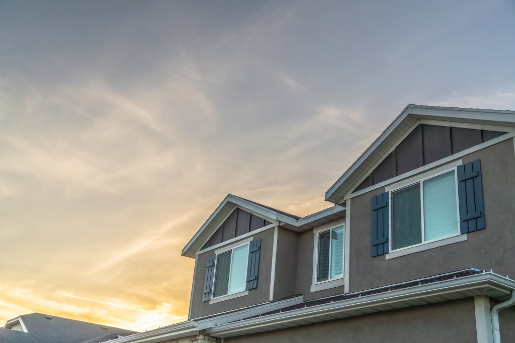 Exterior upper storey of a home with golden cloudy sky background at sunset. The house has gray wall and windows with wooden shutters.