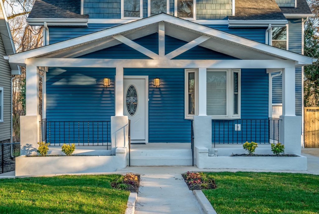 Beautiful renovated craftsman style covered porch with white columns, beams, black baluster railing in front of a blue horizontal vinyl lap siding single family home